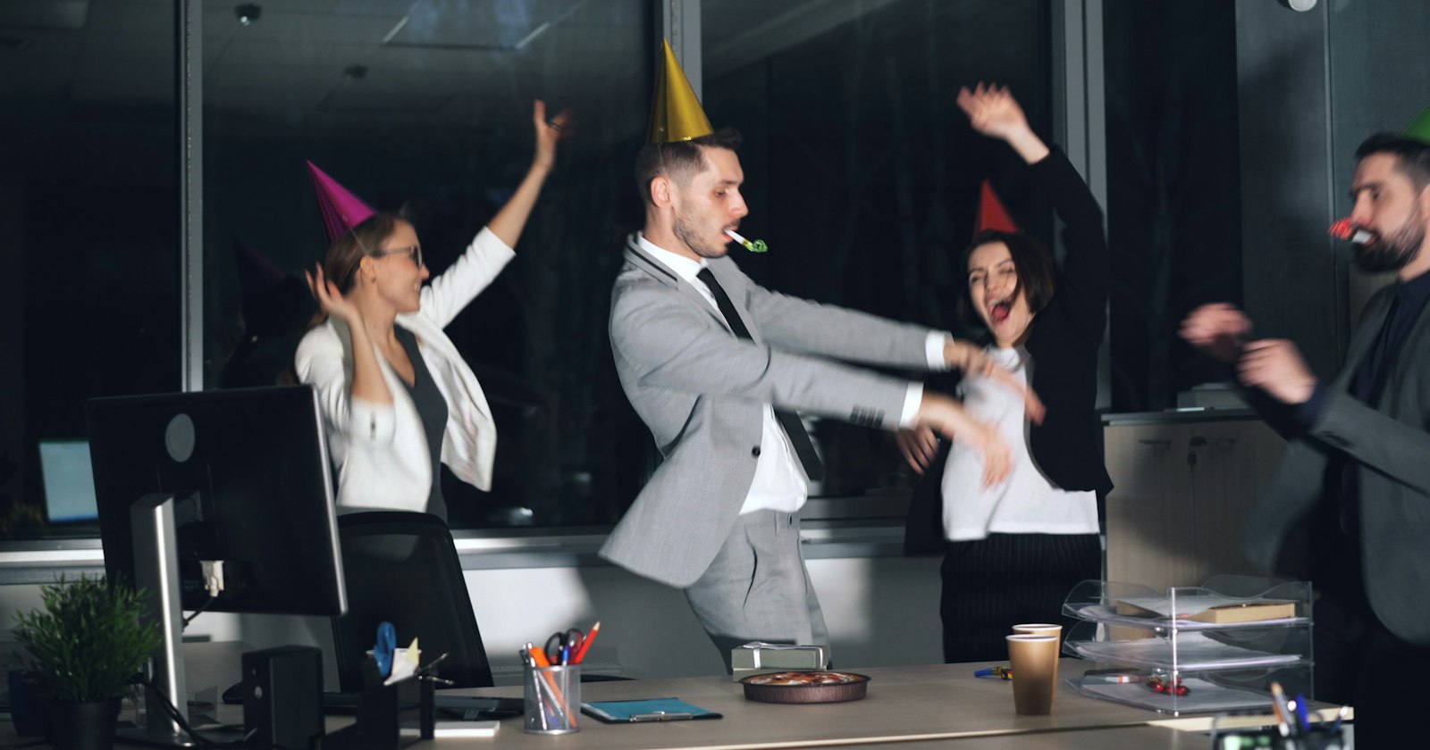 Kolleginnen und Kollegen mit Partyhüten tanzen ausgelassen bei einer Feier im Büro